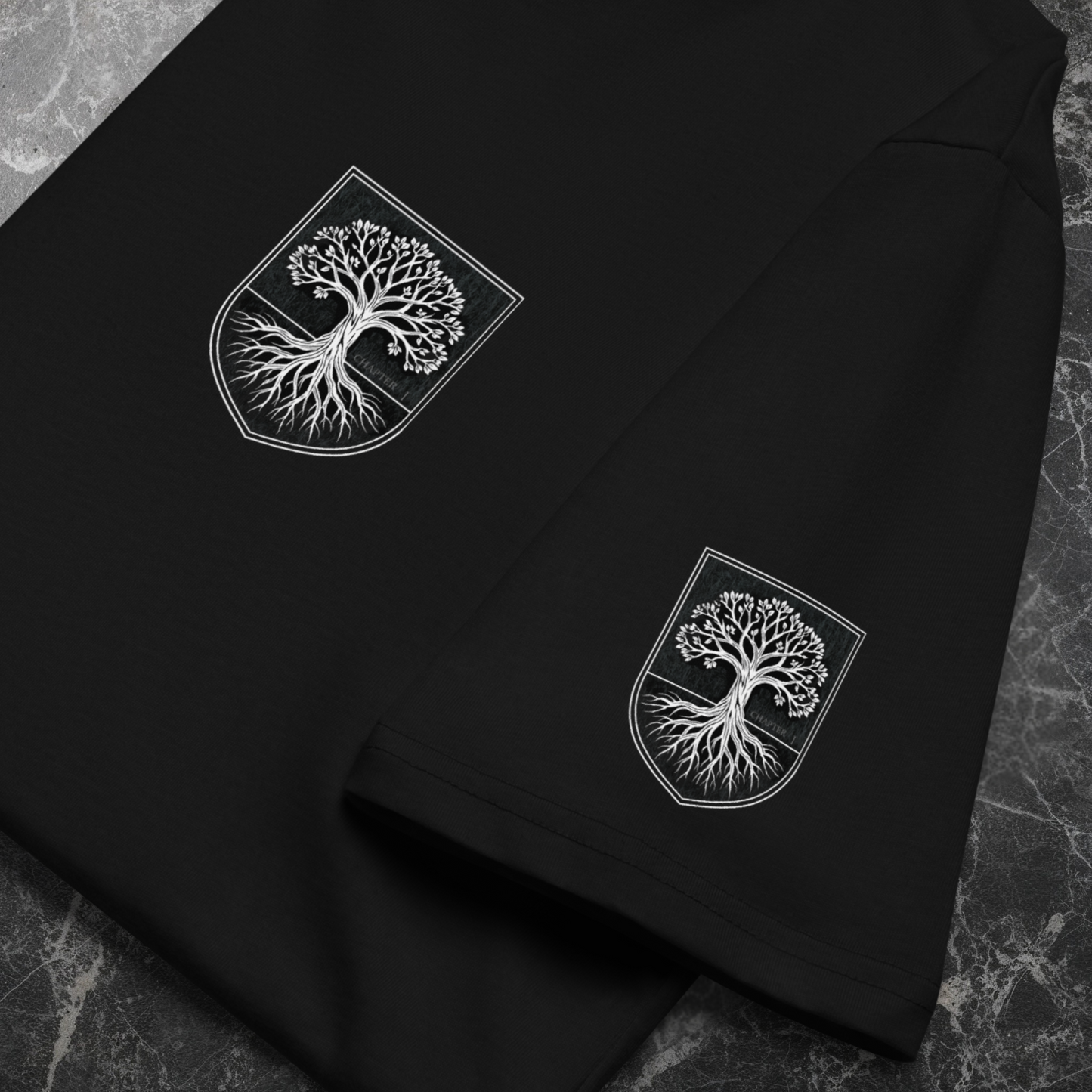 Flat-lay of black organic tee on stone surface; left-chest crest and sleeve crest; sharp DTFlex print detail.