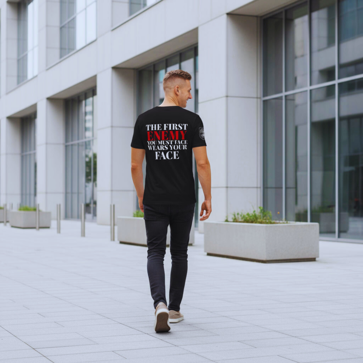 Full-body walk-away in minimalist city setting; black tee back quote visible, ENEMY in red.