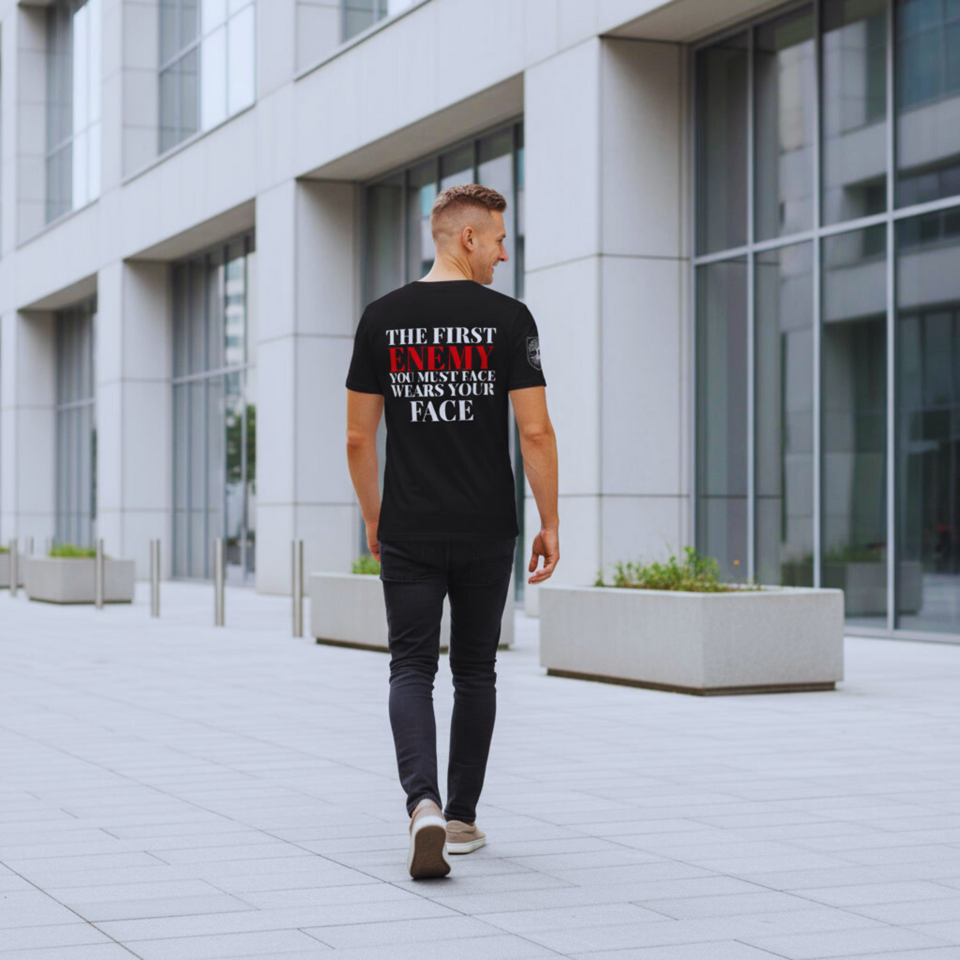 Full-body walk-away in minimalist city setting; black tee back quote visible, ENEMY in red.