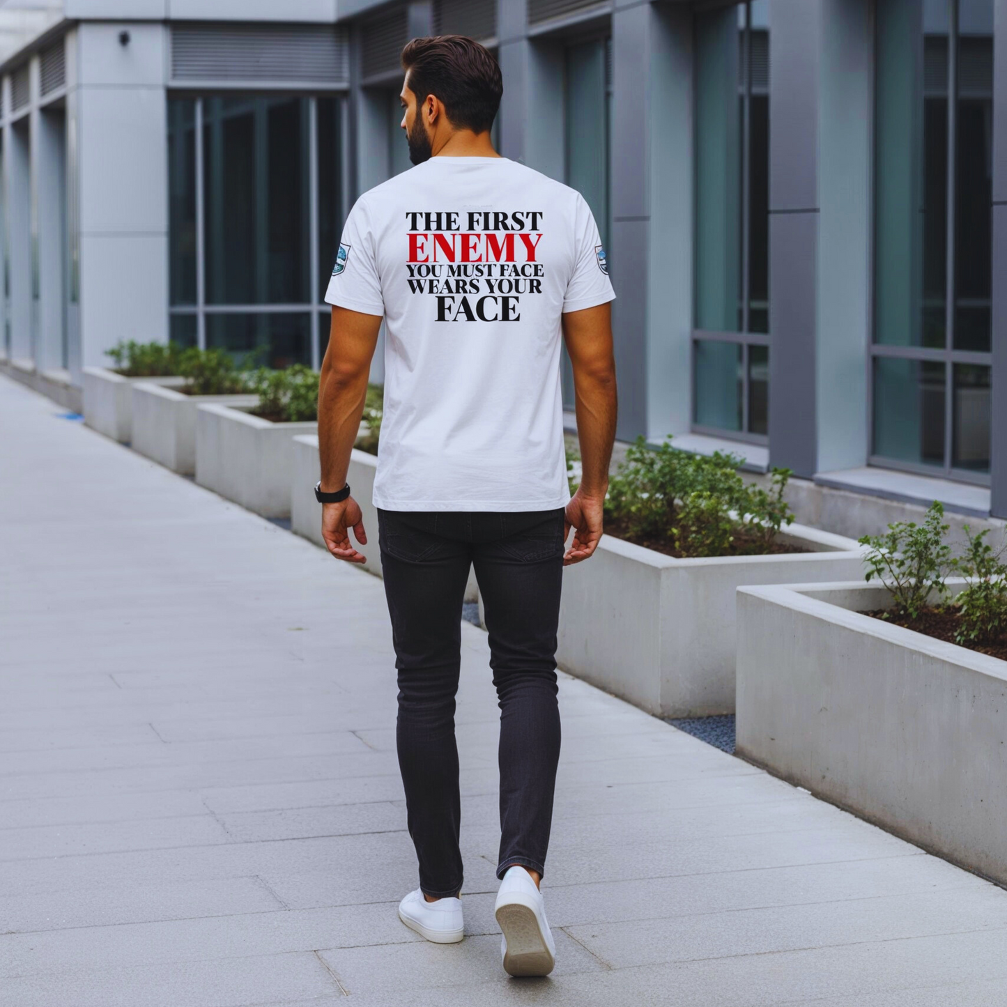 Full-body urban walk-away; white organic tee back quote clearly legible (ENEMY in red), minimalist concrete and glass city setting.