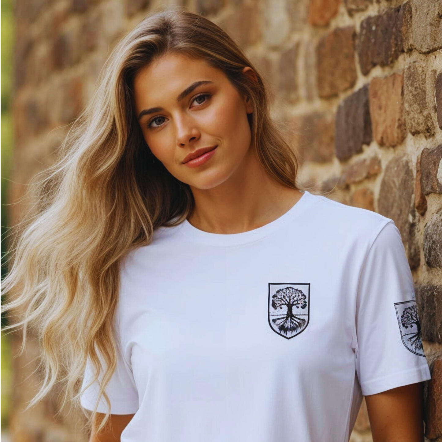 Front view of white organic t-shirt with shield tree-and-roots crest over heart; rustic stone wall background; natural daylight; unisex fit.