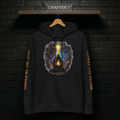 Black “Carry the Flame” hoodie hanging on a wooden hanger against a matte black brick wall under a “Chapter 1” shelf sign, front graphic of the cloaked figure holding a living flame beneath the star fully visible, sleeves hanging straight with the quote text running cleanly down each arm.