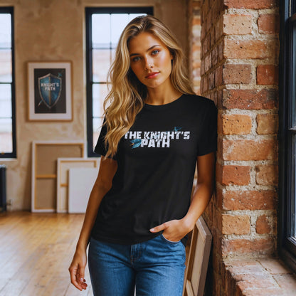 Woman in a loft studio leaning against an exposed brick pillar, wearing a black “The Knight’s Path” organic t-shirt with a distressed white logo and a blue arrow.