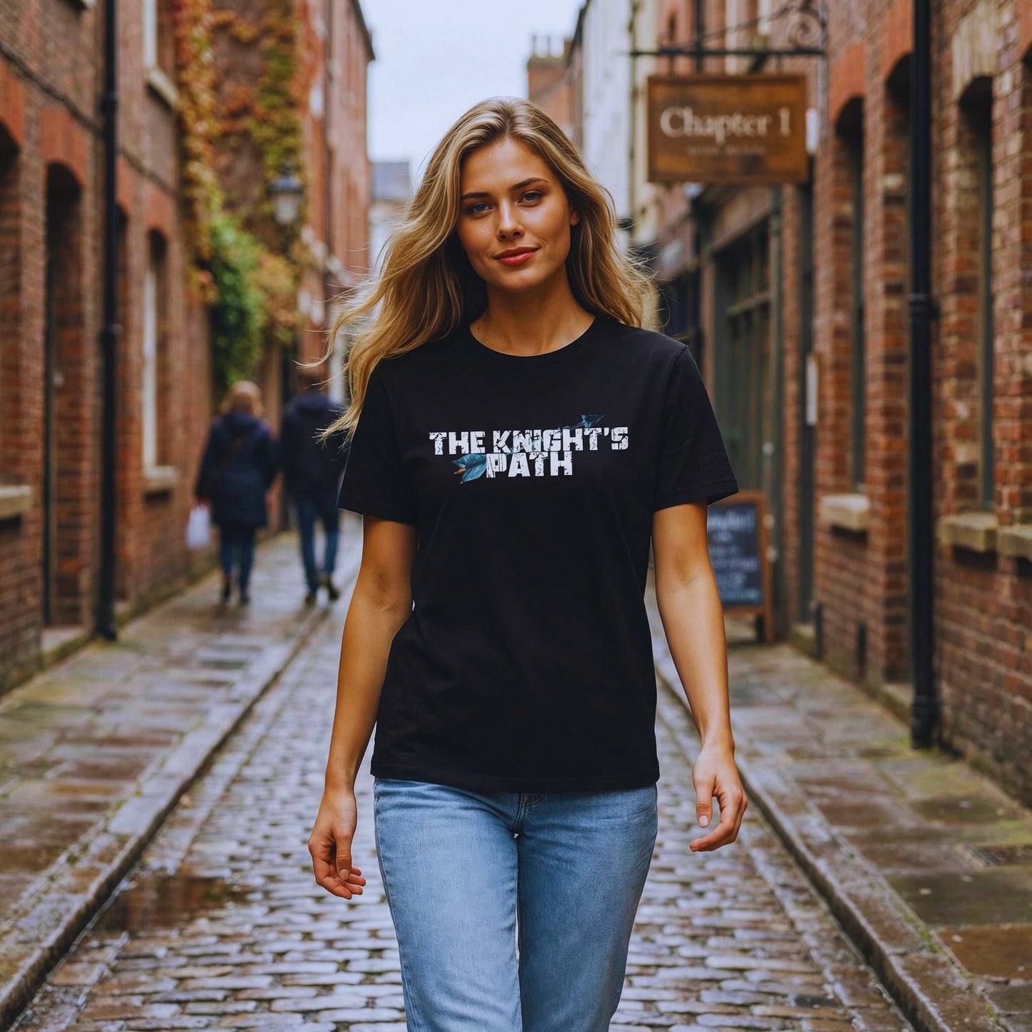 A woman walking down a wet cobblestone alley wearing a black “The Knight’s Path” organic tee with a blue arrow on the chest and a Chapter 1 sign in the background.