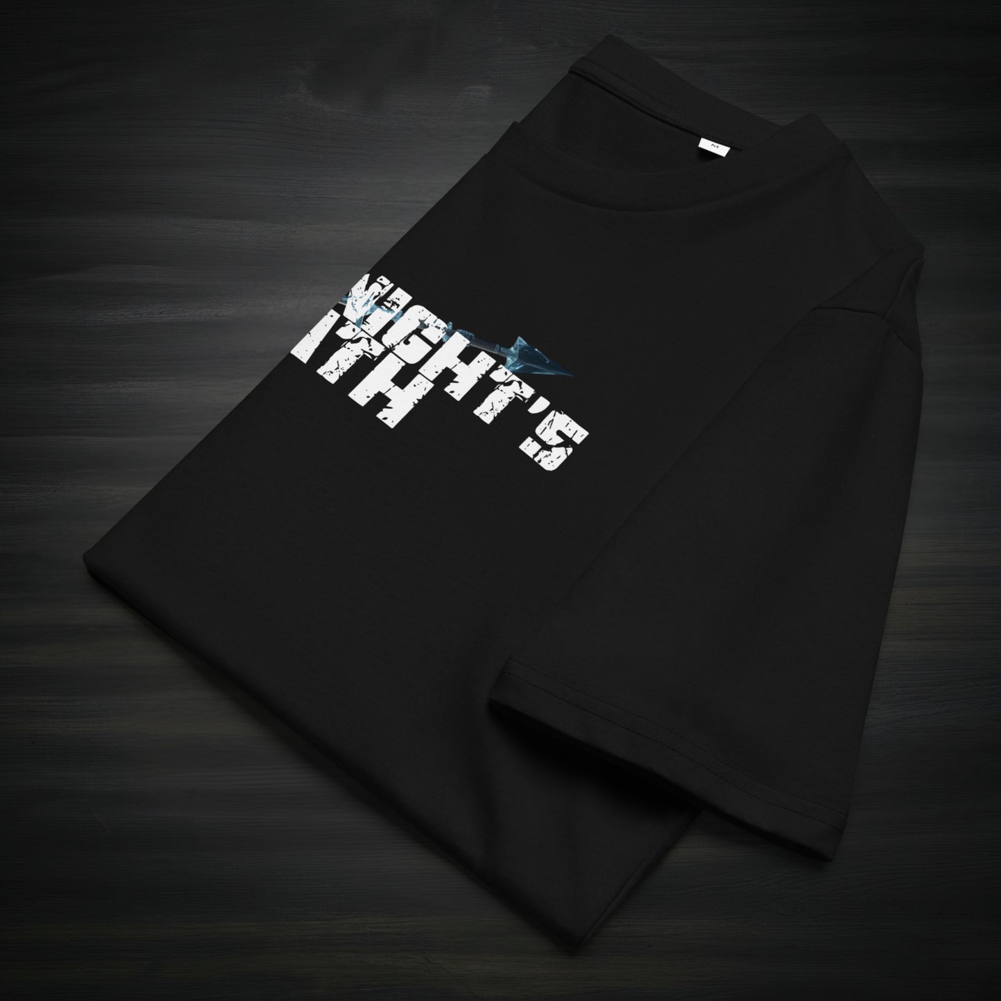 Folded black “The Knight’s Path” organic t-shirt laid on a dark surface, showing the cracked white logo and a blue arrow across the chest.