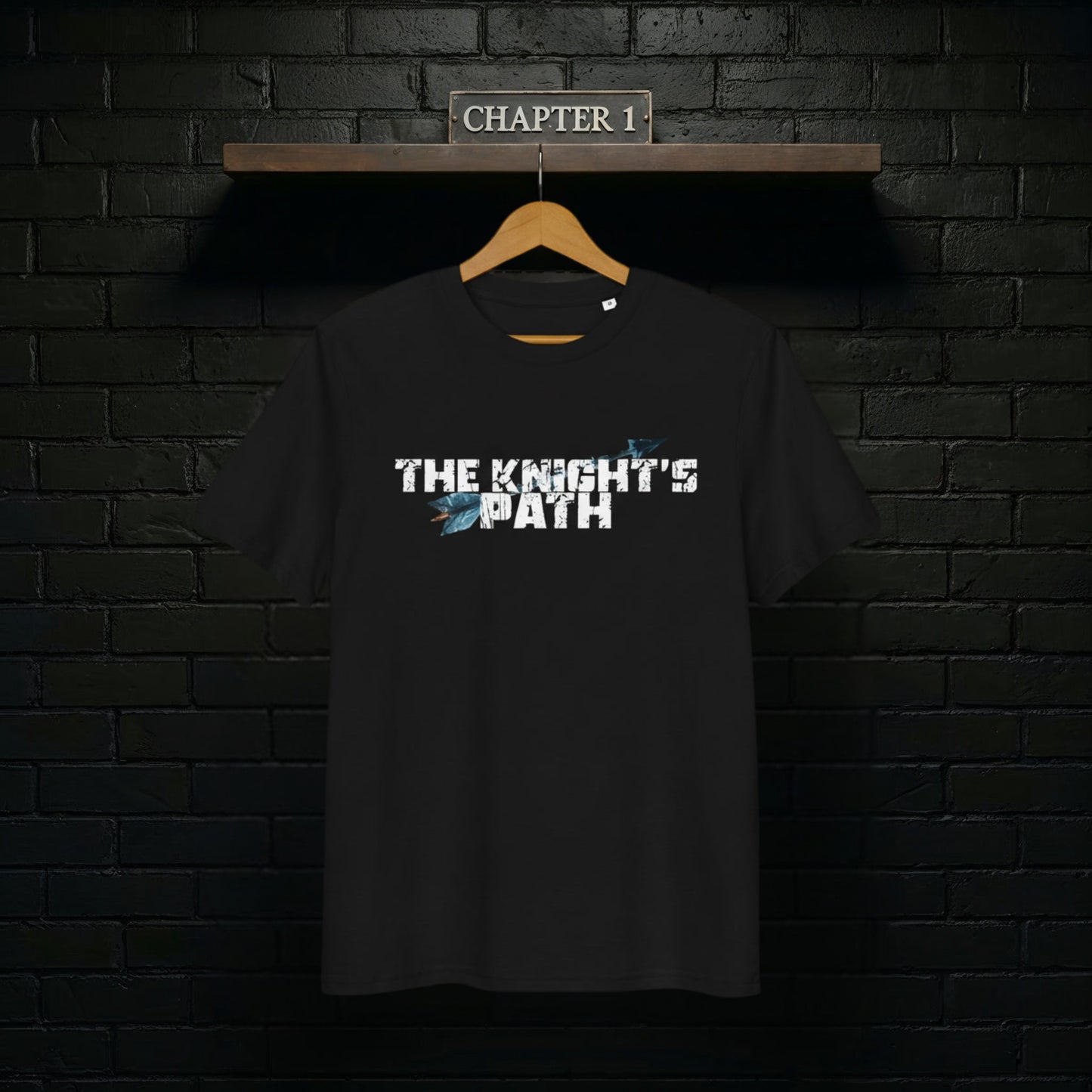 Black “The Knight’s Path” organic tee on a wooden hanger against a black brick wall with a Chapter 1 shelf sign above.