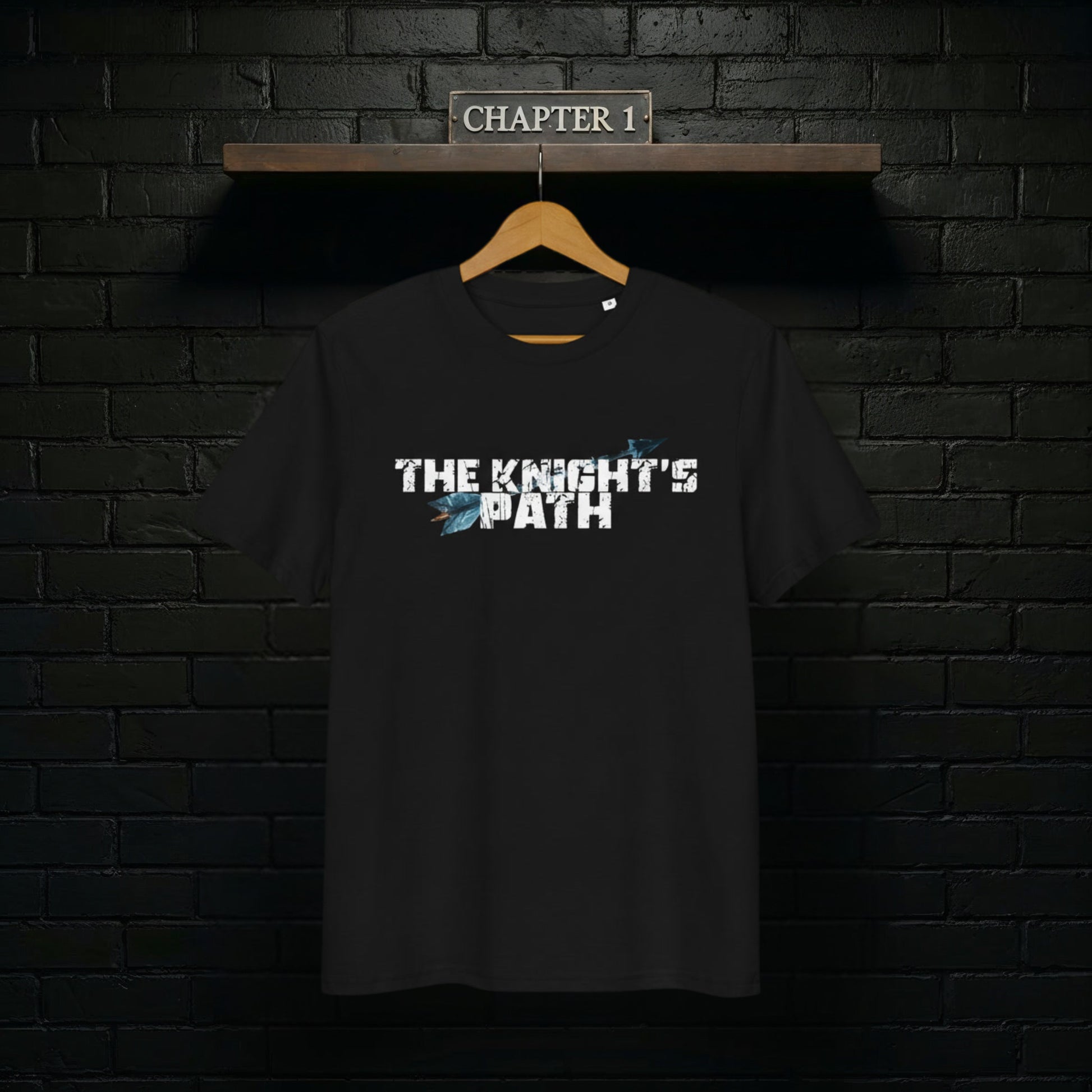 Black “The Knight’s Path” organic tee on a wooden hanger against a black brick wall with a Chapter 1 shelf sign above.