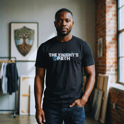 Man standing in an industrial loft wearing a black “The Knight’s Path” organic tee with bold white text and blue arrow, shield artwork blurred in the background.