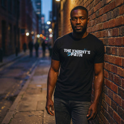 Man in a black “The Knight’s Path” organic t-shirt standing in a moody city alley at dusk, streetlights glowing behind him.