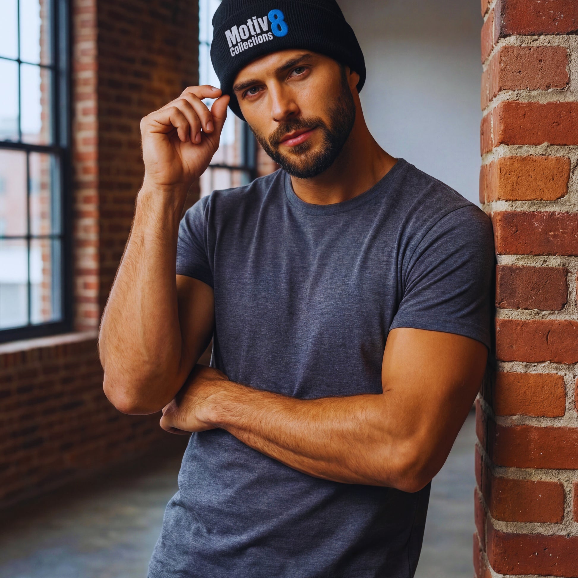 Motiv8 Collections black beanie with blue 8 logo worn by Adrian Holt leaning on a brick pillar in the loft, casual fitted tee and confident pose.