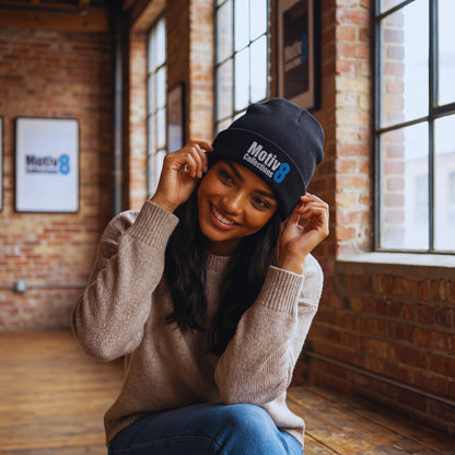 Motiv8 Collections black logo beanie worn by Amara Solis sitting on a wooden bench in the loft, relaxed pose near tall windows and Motiv8 wall art.