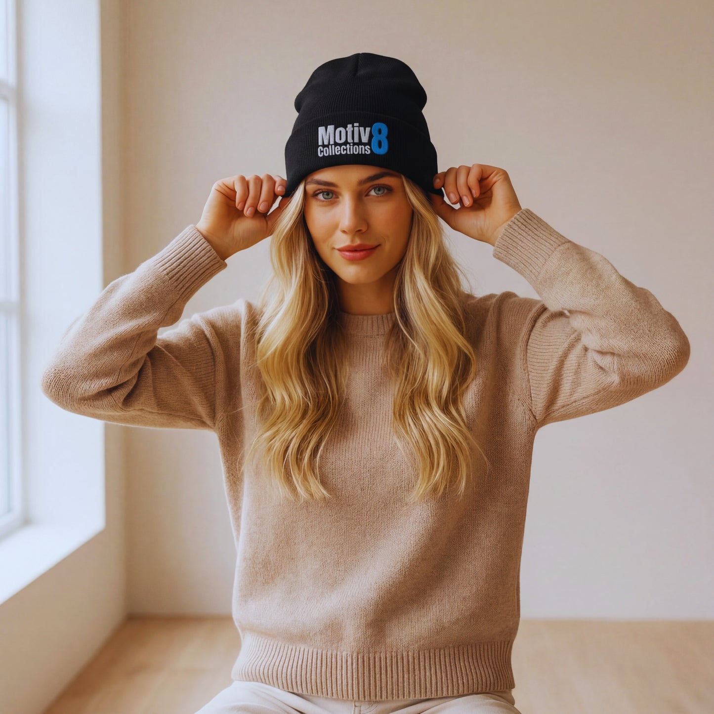 Motiv8 Collections black cuffed beanie with blue 8 logo on Elena Rowan seated against a neutral wall, clean minimal lifestyle styling for the beanie.