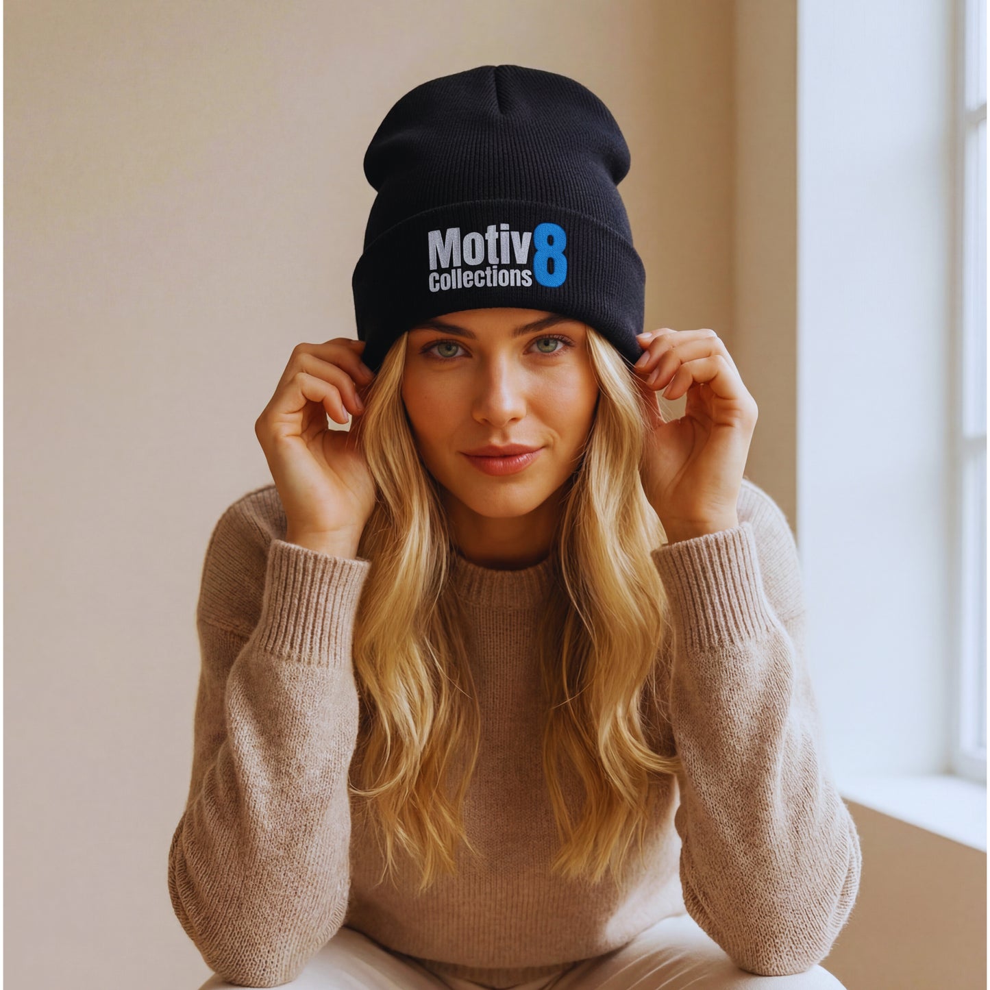 Motiv8 Collections black cuffed beanie with blue 8 logo on Elena Rowan sitting by a bright window, soft natural light and warm knit sweater styling.