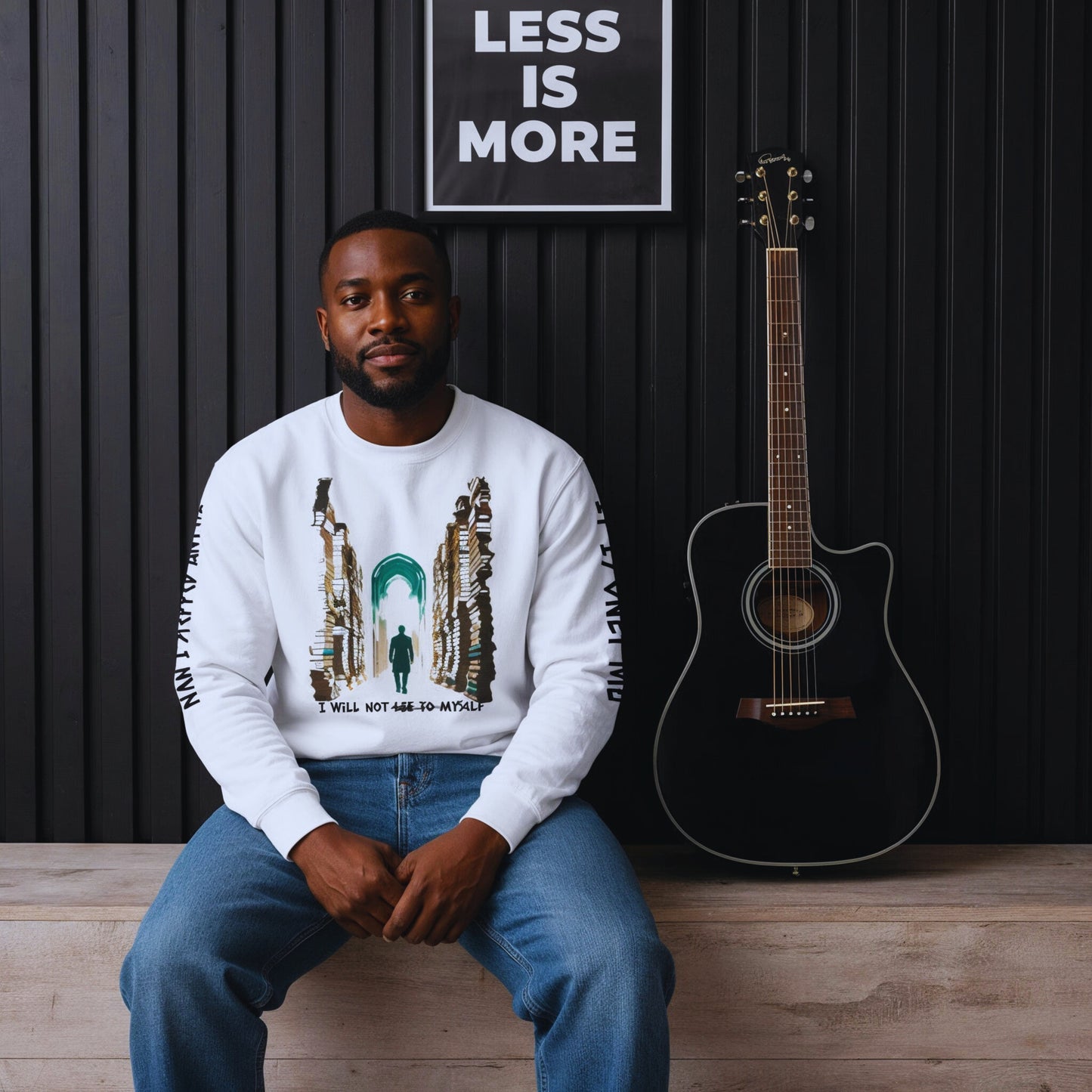 Man in casual studio setting, sitting beside a black guitar under a “Less Is More” poster, wearing the white motivational sweatshirt.