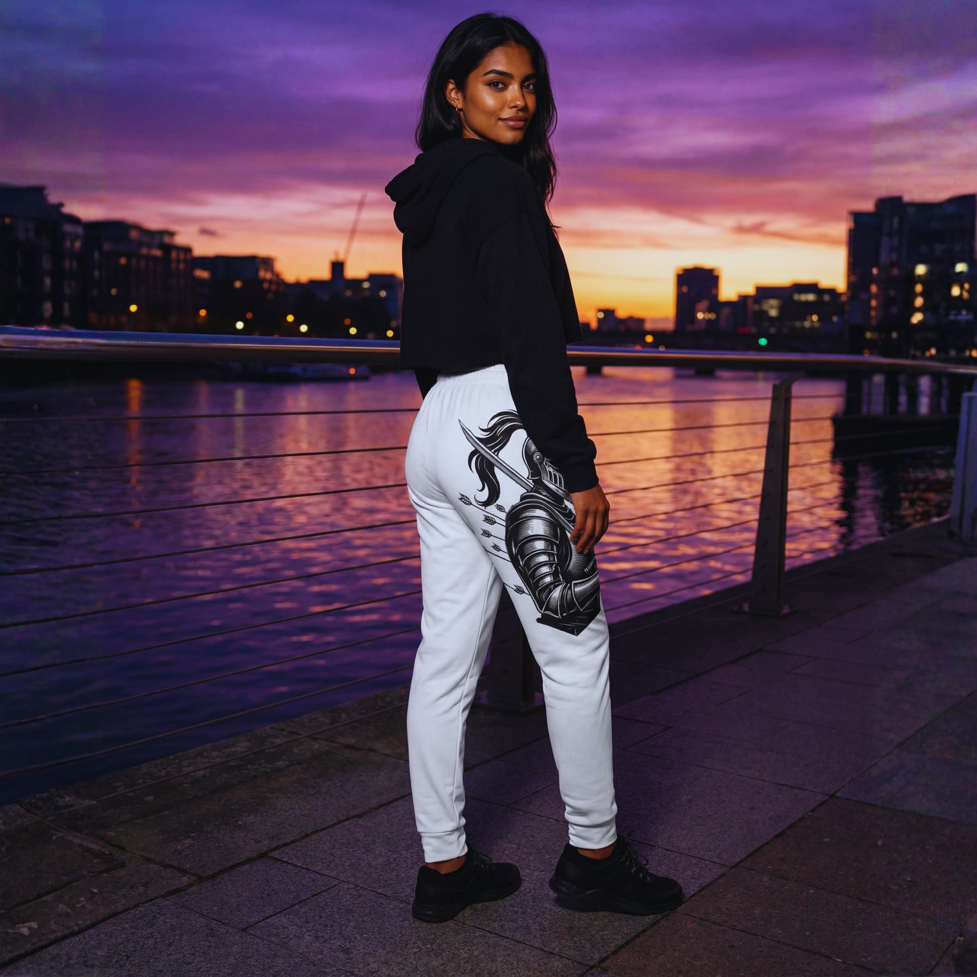 Amara at twilight on a riverside walkway in white Motiv8 Collections Knight Armor joggers and a cropped black hoodie, three-quarter back view with the knight and arrow design glowing against city lights.