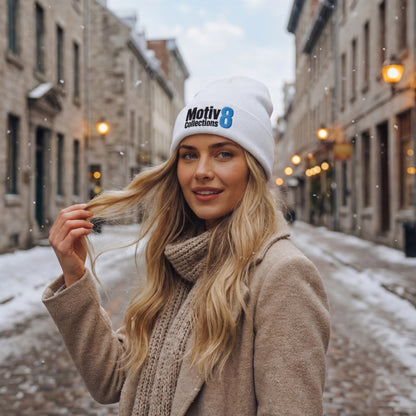 Woman in white Motiv8 Collections beanie and camel coat walking through snowy old town street, cosy winter fashion