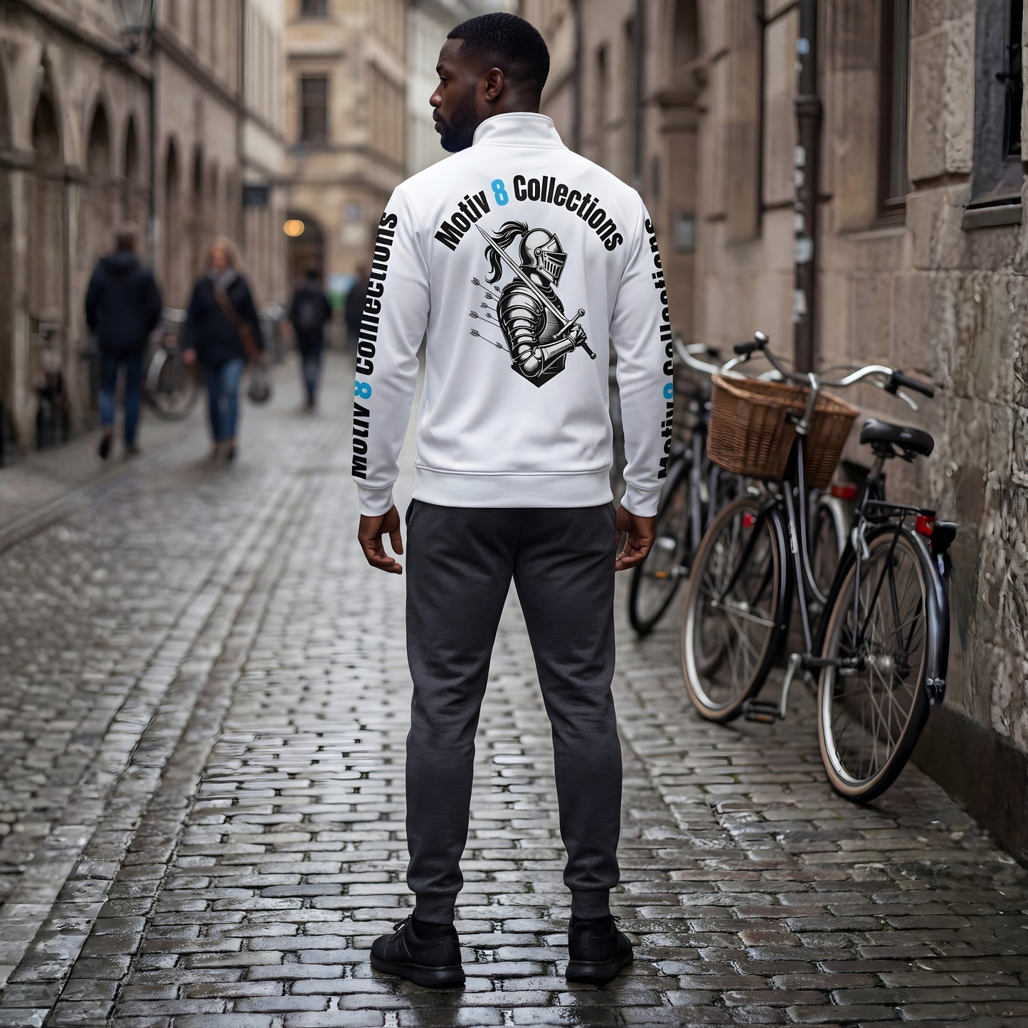 Back view of Malik in the white Motiv8 Collections track jacket on an old-town cobblestone street, clearly displaying the detailed knight illustration with arrows and Motiv8 Collections typography running down both sleeves.