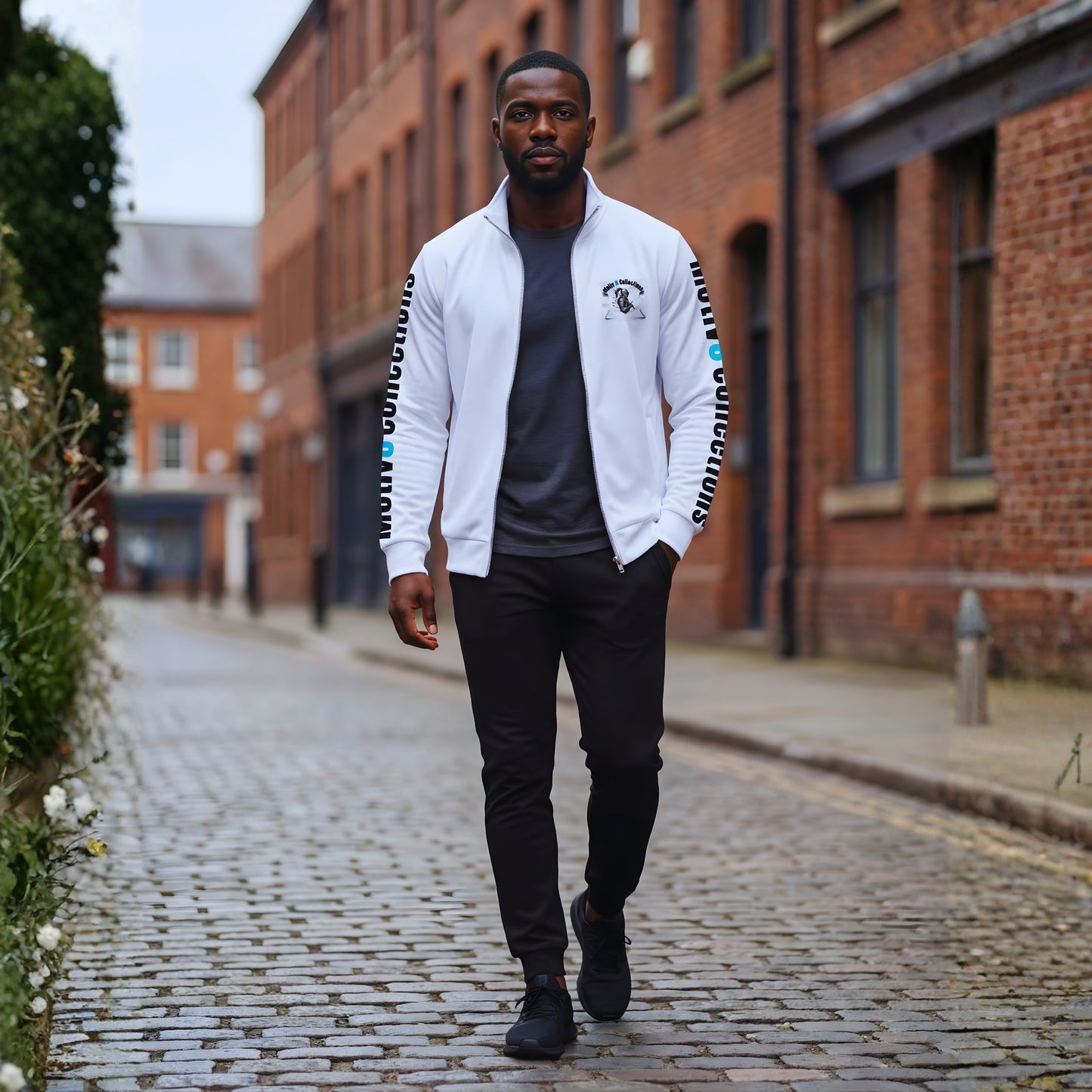 Malik walking along a cobblestone street in the white Motiv8 Collections track jacket, worn open over a dark T-shirt, showing the front logo and sleeve branding in a relaxed urban streetwear scene.