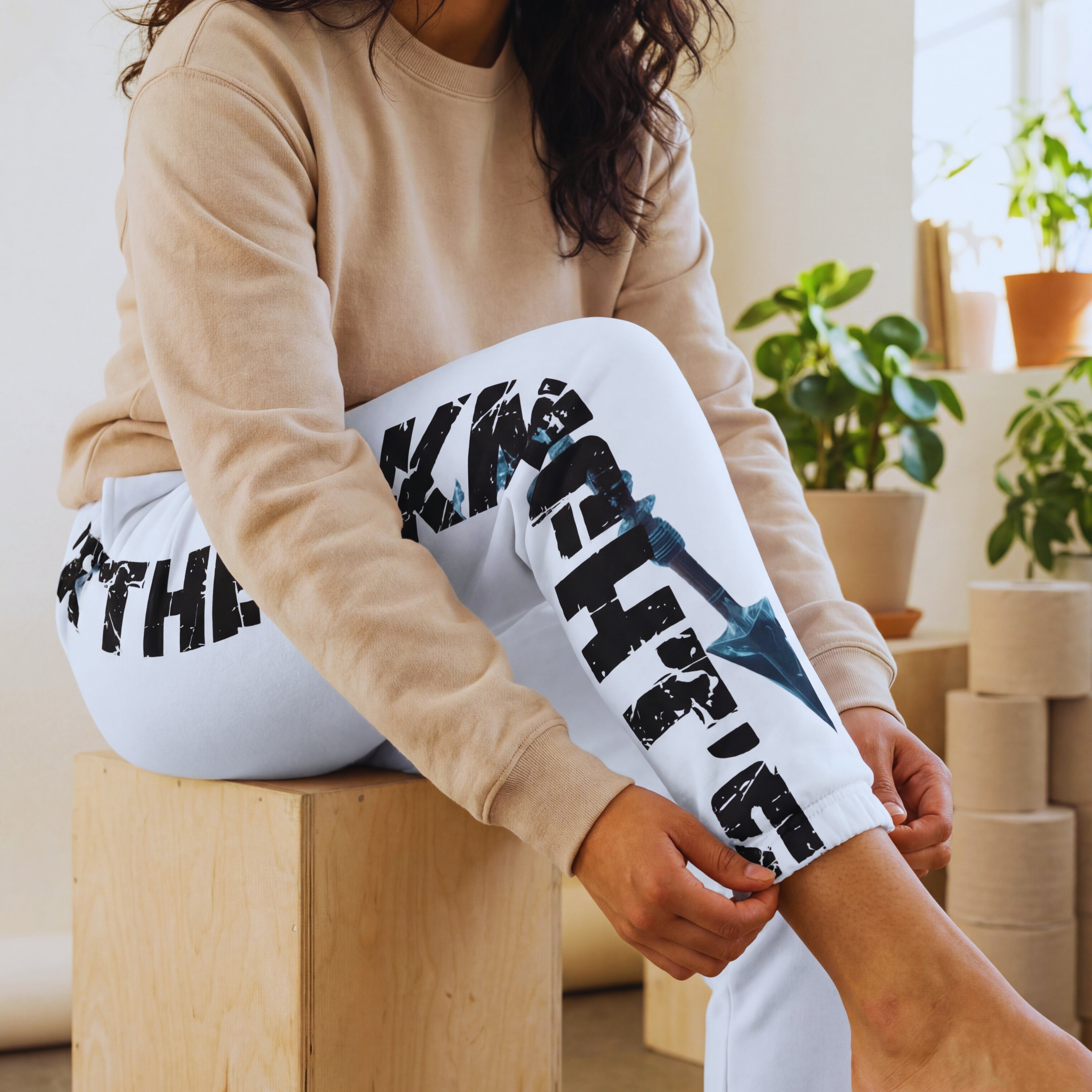 Close-up in eco studio of The Knight’s Path “Arrow One” white joggers, leg bent to highlight distressed “The Knight’s Path” lettering and blue crystal arrow graphic.