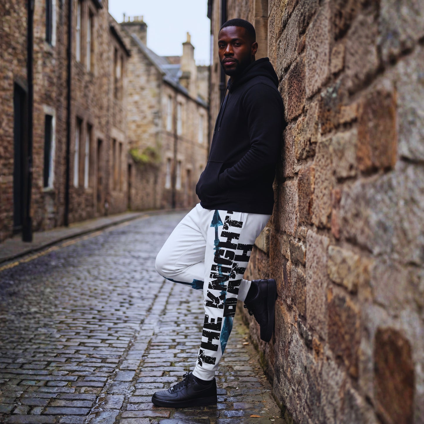 Malik leaning against a stone wall in black hoodie and The Knight’s Path “Arrow One” white joggers, side angle showing the bold arrow and slogan down the leg.