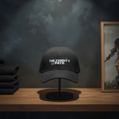 Black “The Knight’s Path” arrow logo cap on a display stand in a moody fantasy shop, framed by folded tees and warrior artwork behind.
